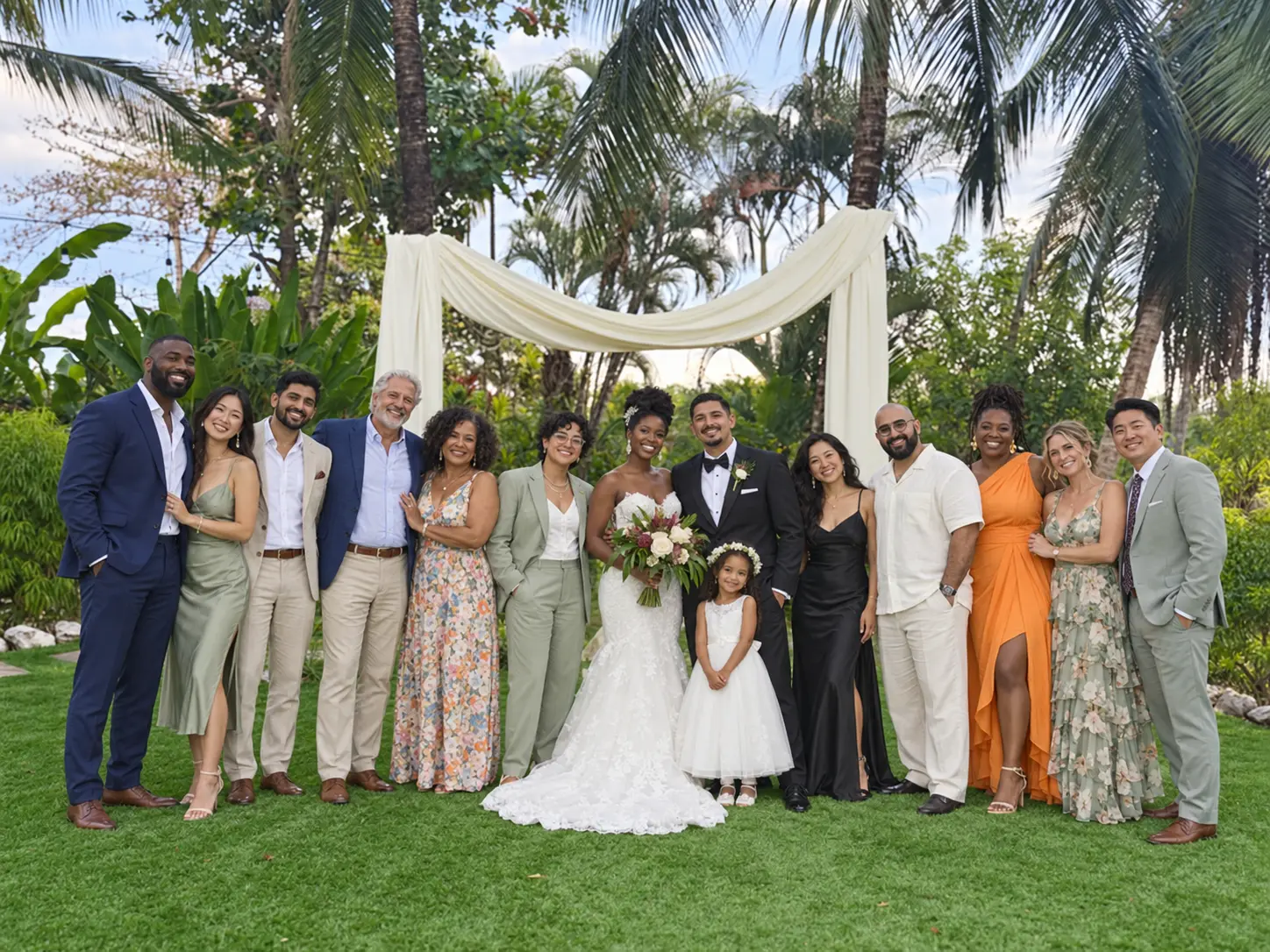 A newlywed couple with their families on a garden lawn beneath a draped ceremony arch