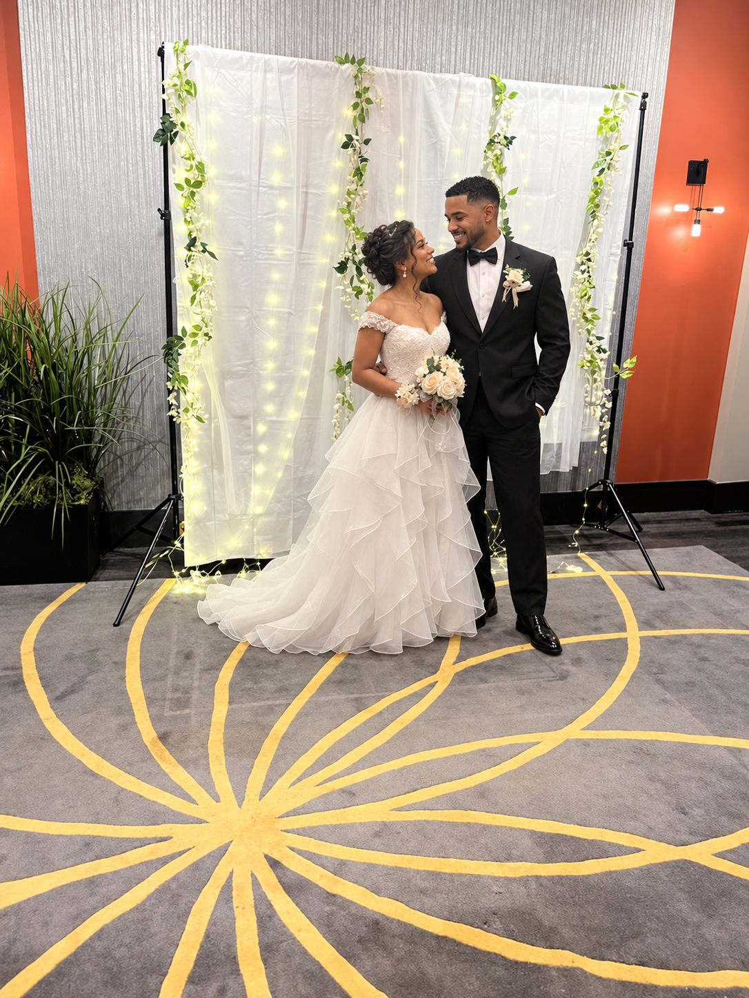 Newlyweds smiling at each other in front of a floral ceremony backdrop with string lights